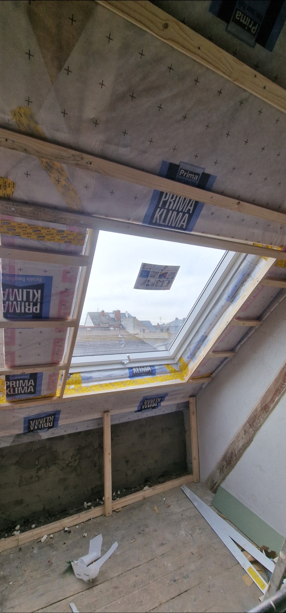 Montaż okna dachowego w skośnym dachu, widoczne drewniane ramy, folia paroizolacyjna z nadrukiem, fragment poddasza w trakcie prac wykończeniowych.