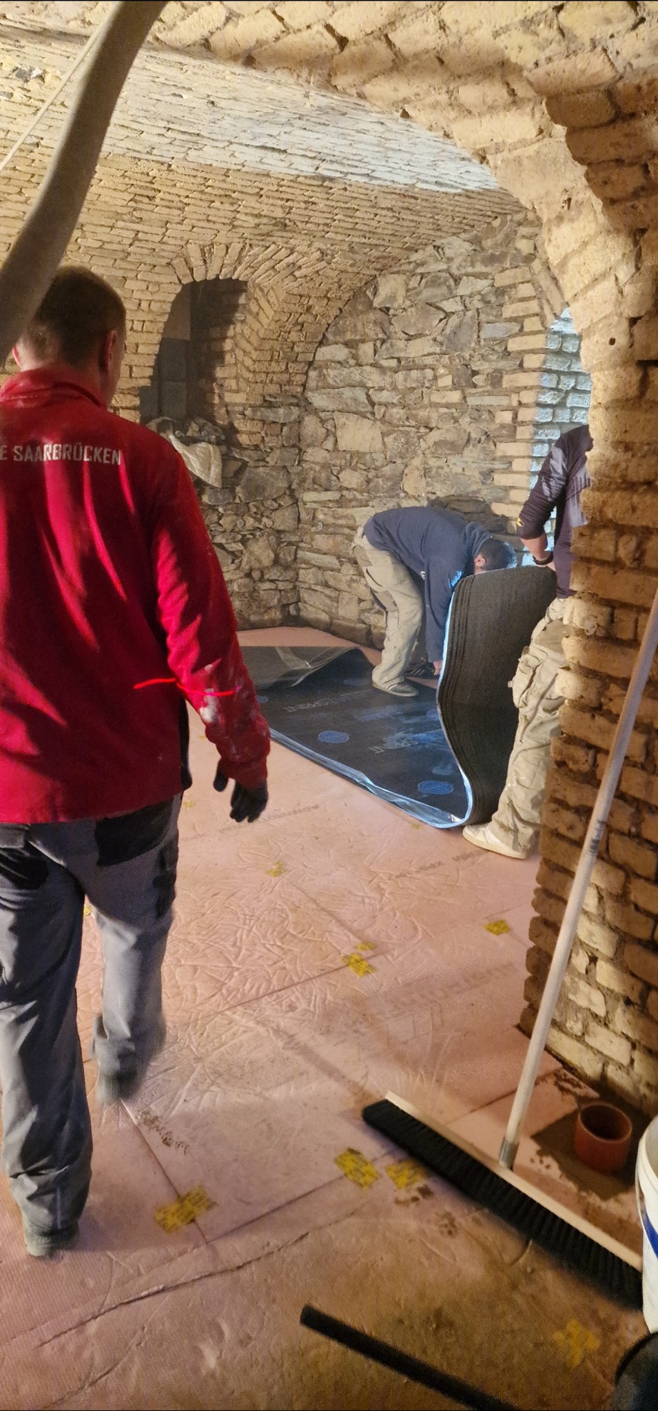 Prace izolacyjne w piwnicy ze sklepieniem łukowym: ekipa montuje czarną matę izolacyjną na różowej warstwie styropianu, widoczne żółte elementy mocujące.