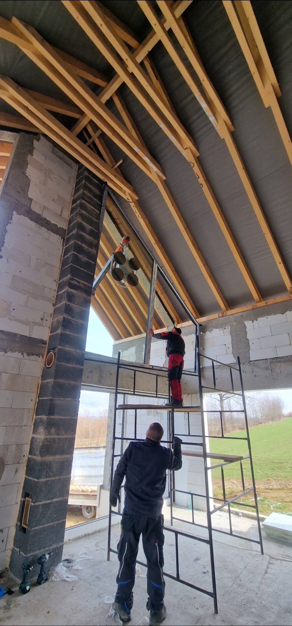 Montaż okna dachowego na rusztowaniu w surowym wnętrzu. Widoczne drewniane elementy konstrukcji dachu i narzędzia. Praca na wysokości, jasne światło dzienne.
