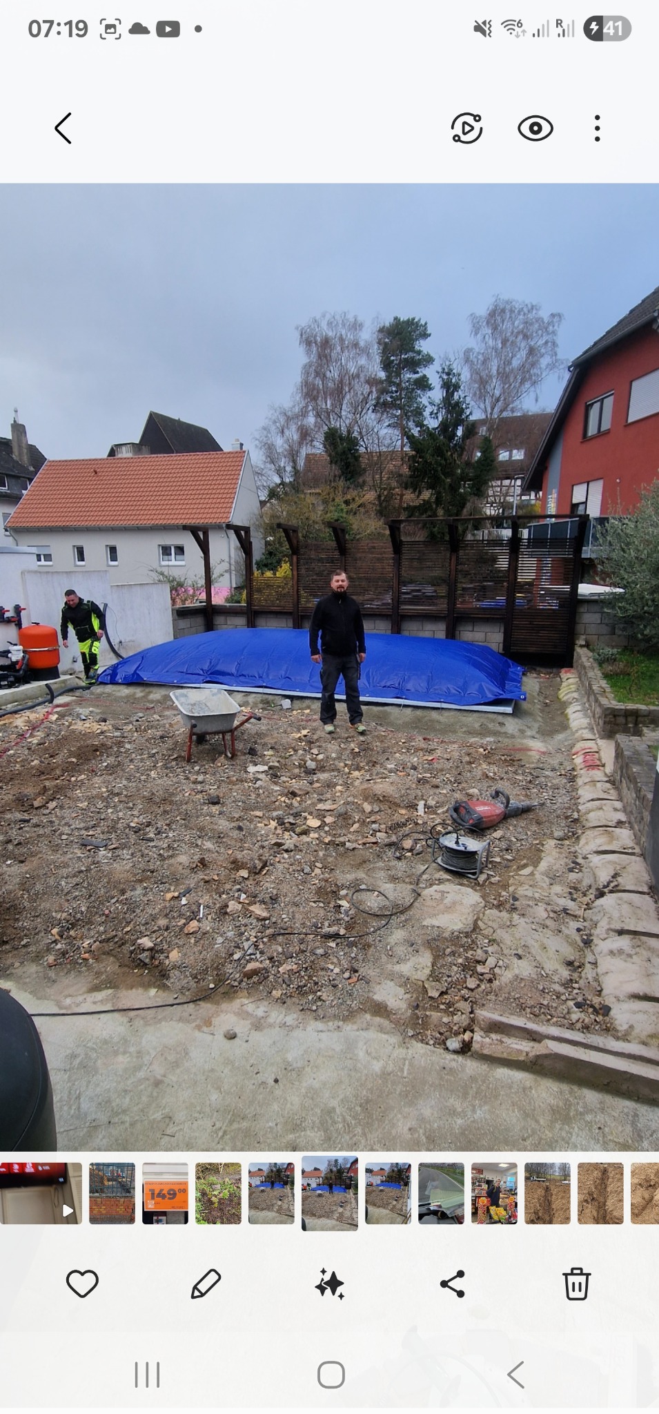 Przygotowanie terenu pod basen: robotnicy, narzędzia (wózek, młot pneumatyczny), zabezpieczona niecka basenowa plandeką. Widok na domy w tle.