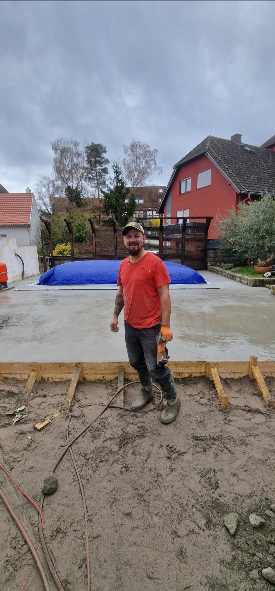 Uśmiechnięty mężczyzna w roboczych butach stoi na świeżo wylanej betonowej płycie, otoczonej drewnianym szalunkiem. W tle dom i przykryty basen.