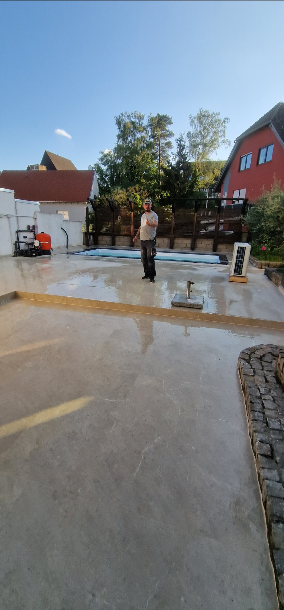 Świeżo wylana, gładka posadzka betonowa na zewnątrz, z widocznym basenem i instalacją. Fachowiec w tle, w słoneczny dzień.