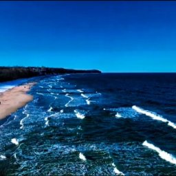 TwoSight Photography - Zimowy krajobraz nadmorski: błękitne morze z falami, piaszczysta plaża pokryta śniegiem, spacerujący ludzie i las w tle pod bezchmurnym niebem. Ujęcie z lotu ptaka.