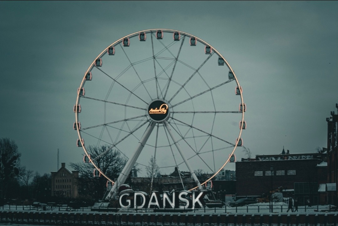 Diabelski młyn w Gdańsku z napisem 'GDAŃSK' na pierwszym planie, sfotografowany w pochmurny dzień. Oświetlone kabiny i logo 'AmberSky' na kole.