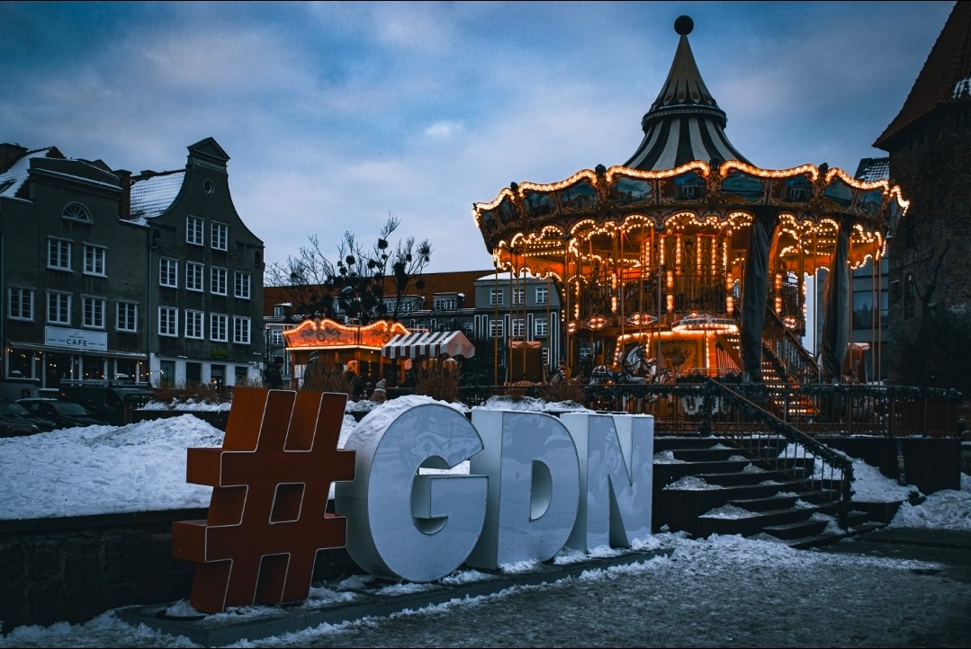 Zimowy krajobraz Gdańska z karuzelą w tle, oświetloną ciepłym światłem. Na pierwszym planie duży, przestrzenny napis #GDN w śniegu, z budynkami w tle.