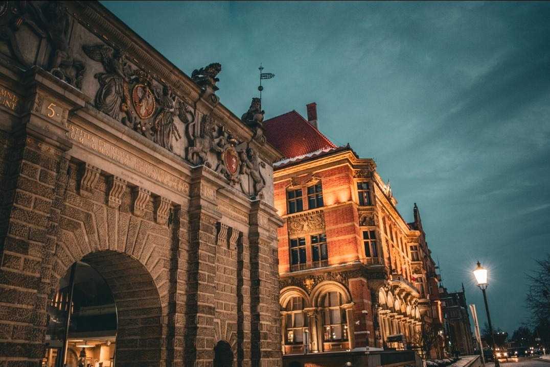 Nocne ujęcie ceglanej bramy z herbem i zdobieniami, obok oświetlonego budynku z czerwonym dachem. Lampa uliczna w tle, pochmurne niebo.