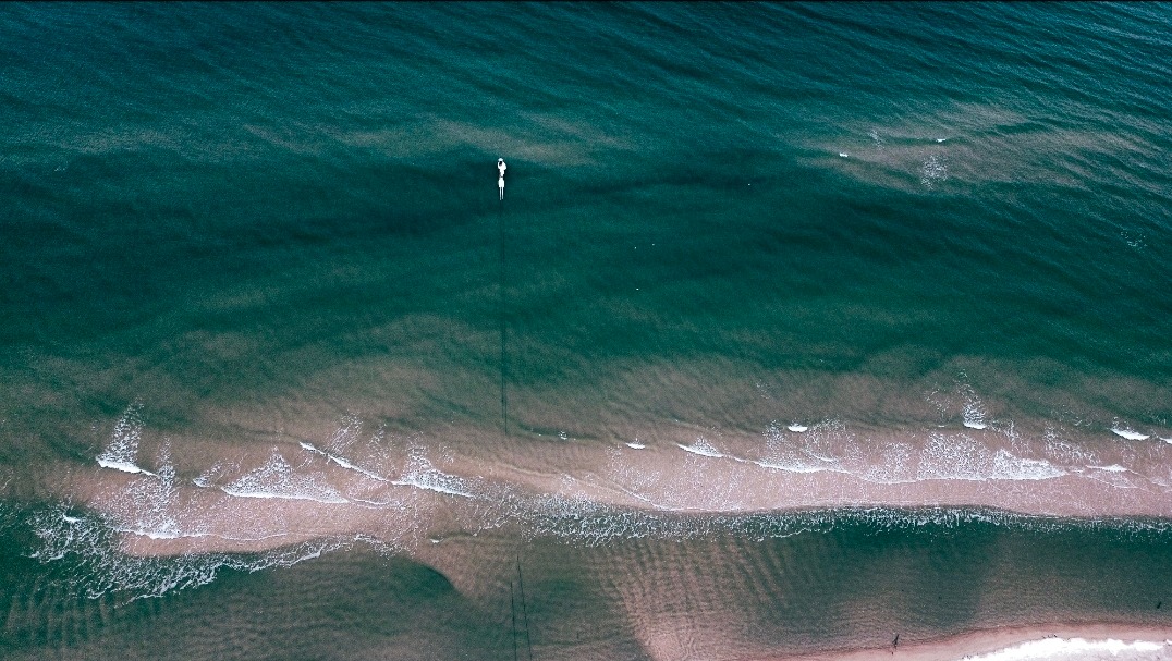 Widok z góry na turkusowe morze z piaszczystą plażą i falami. Dwie osoby na desce SUP płyną po spokojnej wodzie, tworząc minimalistyczny krajobraz.