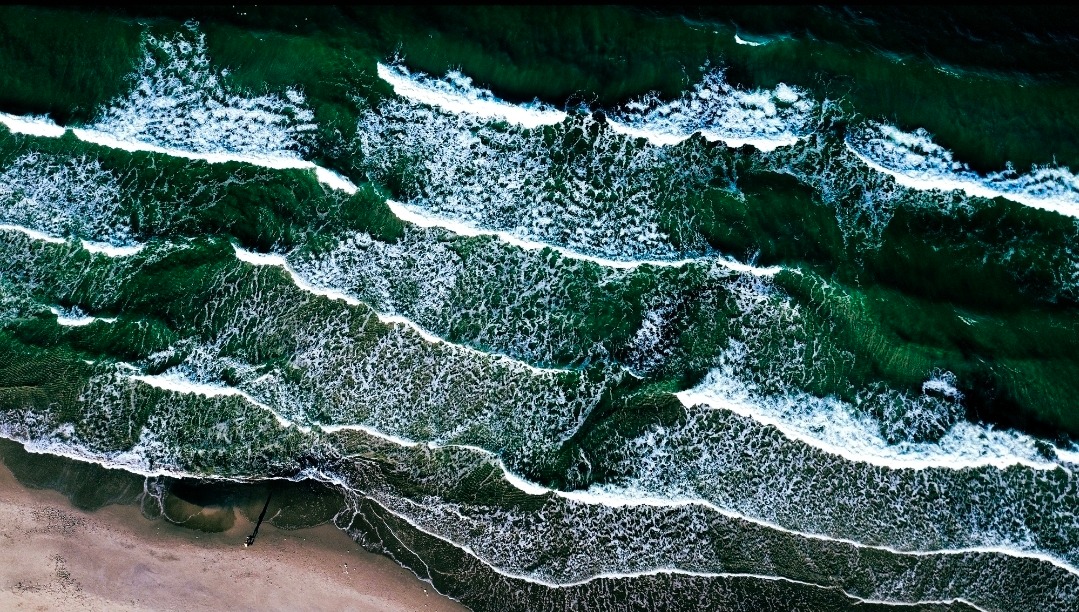 Zdjęcie z lotu ptaka: ciemnozielone fale oceanu z białymi grzywami spienionej wody uderzają o piaszczystą plażę. Widok z góry, kontrastowe kolory, dynamiczne ujęcie.
