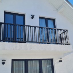 METAL PROCESSING KATARZYNA PROKOP-DZIEDZIC - Czarna, metalowa balustrada balkonowa z pionowymi szczeblami, zamontowana na białym budynku z czarnymi oknami. Widok z dołu, oświetlenie dzienne.