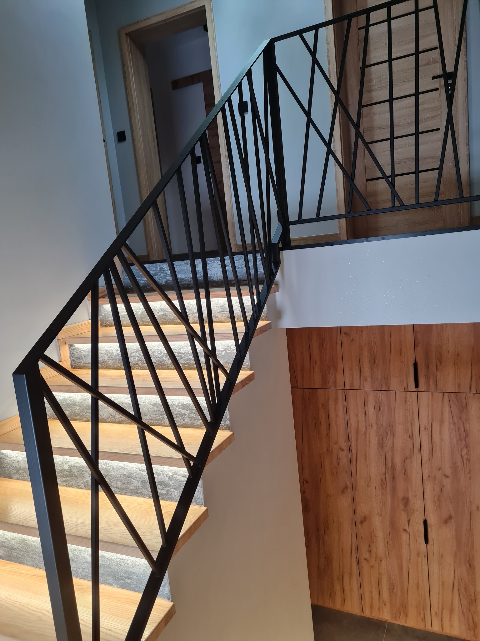 Nowoczesne schody z podświetleniem LED i czarną, geometryczną balustradą. Widoczne drewniane stopnie i drzwi z motywem kratki w tle. Minimalistyczny design.