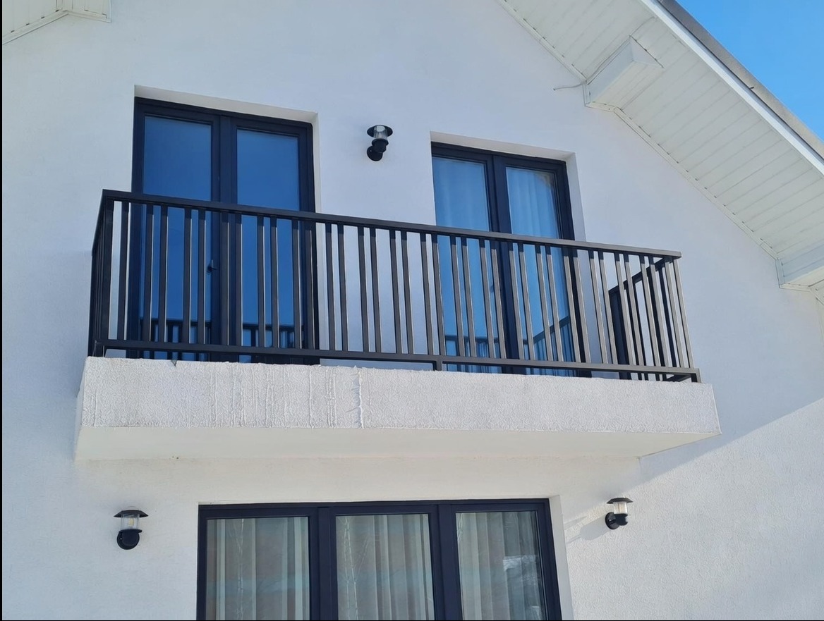 Czarna, metalowa balustrada balkonowa z pionowymi szczeblami, zamontowana na białym budynku z czarnymi oknami. Widok z dołu, oświetlenie dzienne.