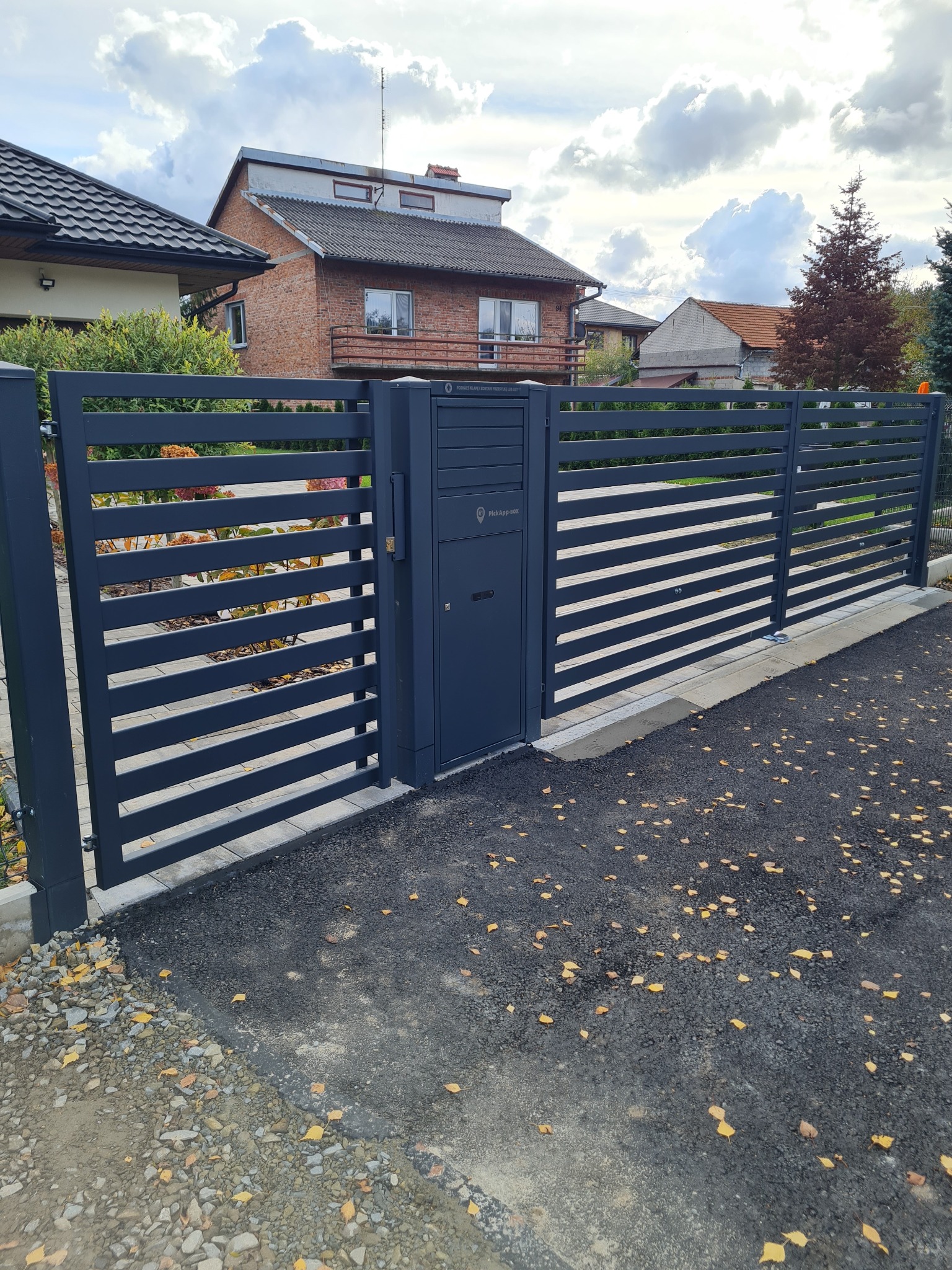 Nowoczesna, pozioma brama wjazdowa z furtką i skrzynką na listy, w kolorze grafitowym, na tle domów jednorodzinnych i jesiennych liści na asfalcie.