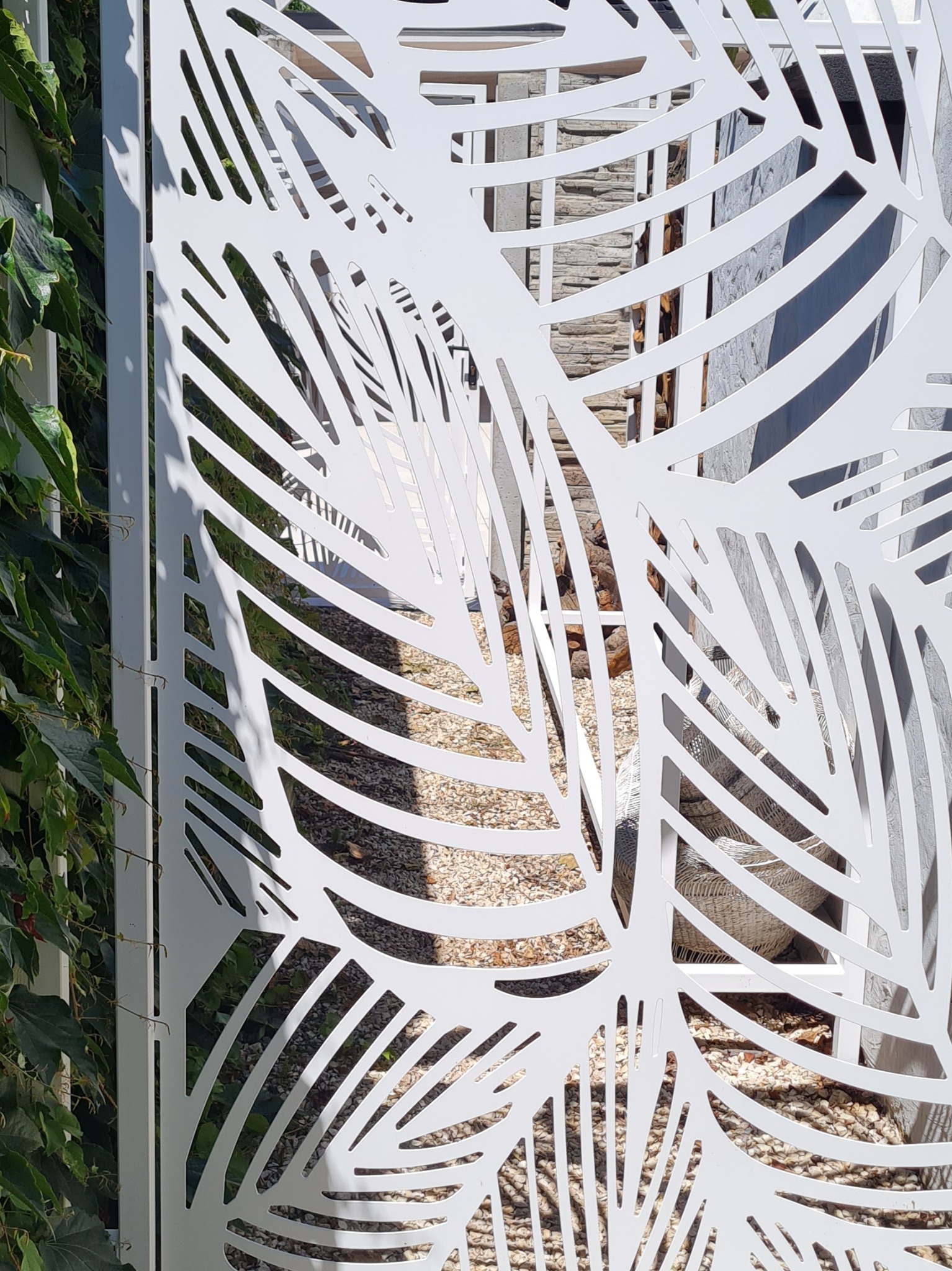 Białe, metalowe ogrodzenie z ażurowym wzorem liści na tle zieleni i żwirowej nawierzchni. Nowoczesny design, precyzyjne wykonanie, jasne oświetlenie.