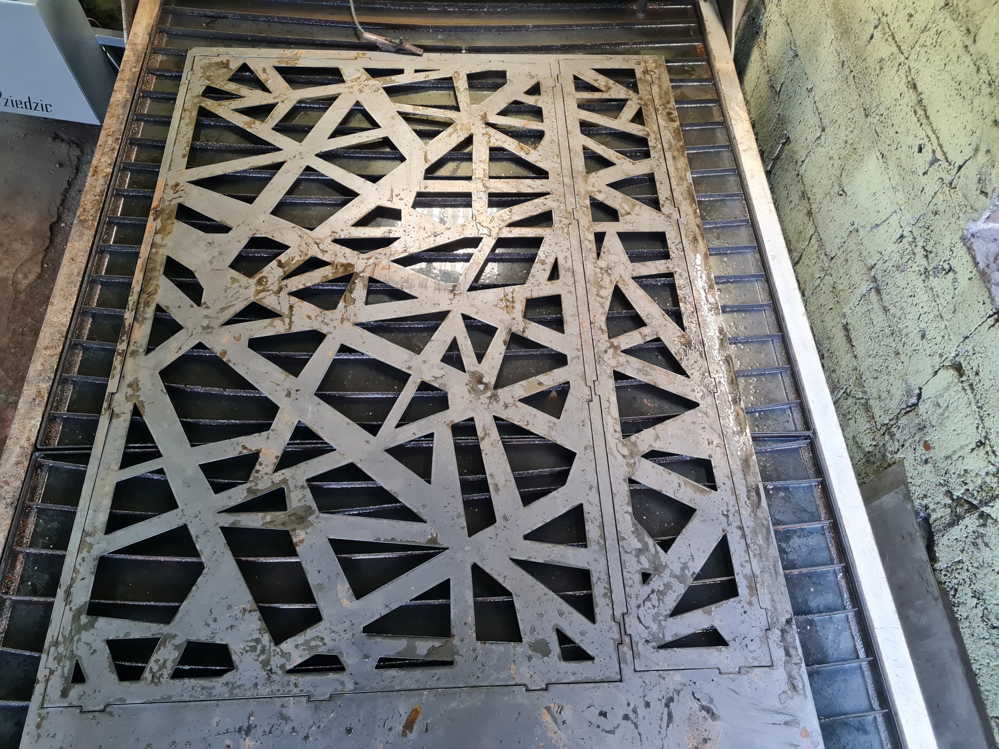 Metalowy panel z wyciętym wzorem geometrycznym, prawdopodobnie po obróbce CNC, leżący na metalowej konstrukcji z widocznymi śladami cieczy chłodzącej.