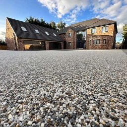 Zywice.pro - Nowoczesny dom z podjazdem wyłożonym drobnym, jasnym kamieniem. Ujęcie z niskiej perspektywy podkreśla fakturę kamienia i bryłę budynku.