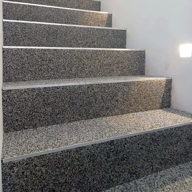 Wykończone schody z szarego, drobnoziarnistego kamienia z metalowymi listwami krawędziowymi, oświetlone kwadratową lampą ścienną.