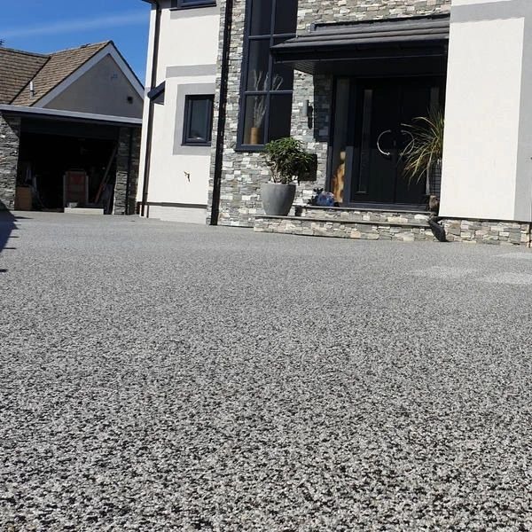 Szary, żywiczny taras przed domem z kamienną elewacją i ciemnymi drzwiami wejściowymi. Donice z roślinami po bokach wejścia. Nowoczesny styl.