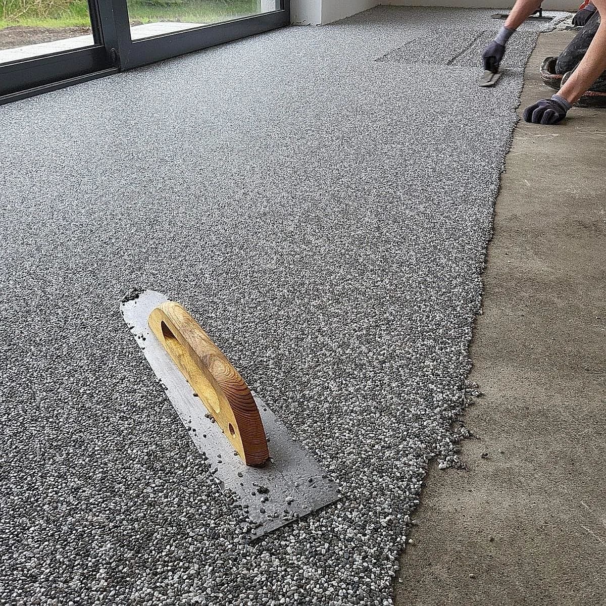 Aplikacja żywicy kamiennej na tarasie z użyciem pacy. Widoczne wykończone fragmenty i surowy beton. Przesuwne drzwi balkonowe w tle.