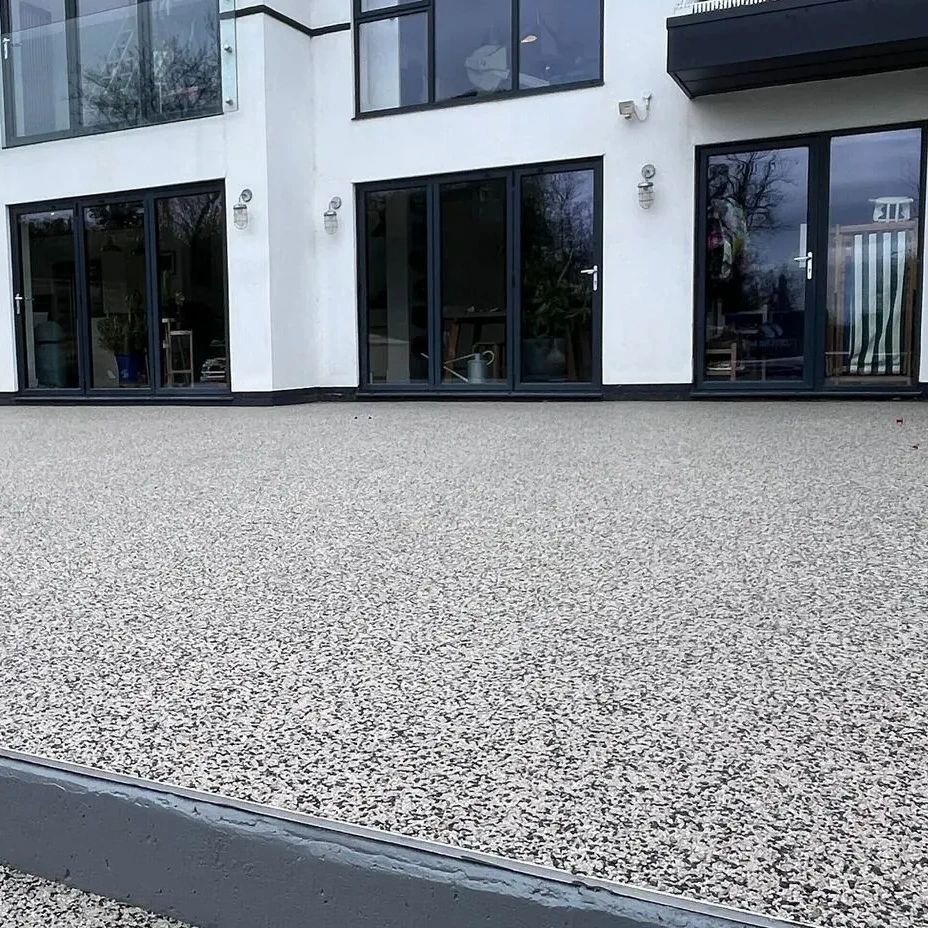 Nowoczesny taras z żywicy epoksydowej w odcieniach szarości, przylegający do białego budynku z dużymi, przeszklonymi drzwiami. Minimalistyczny design.