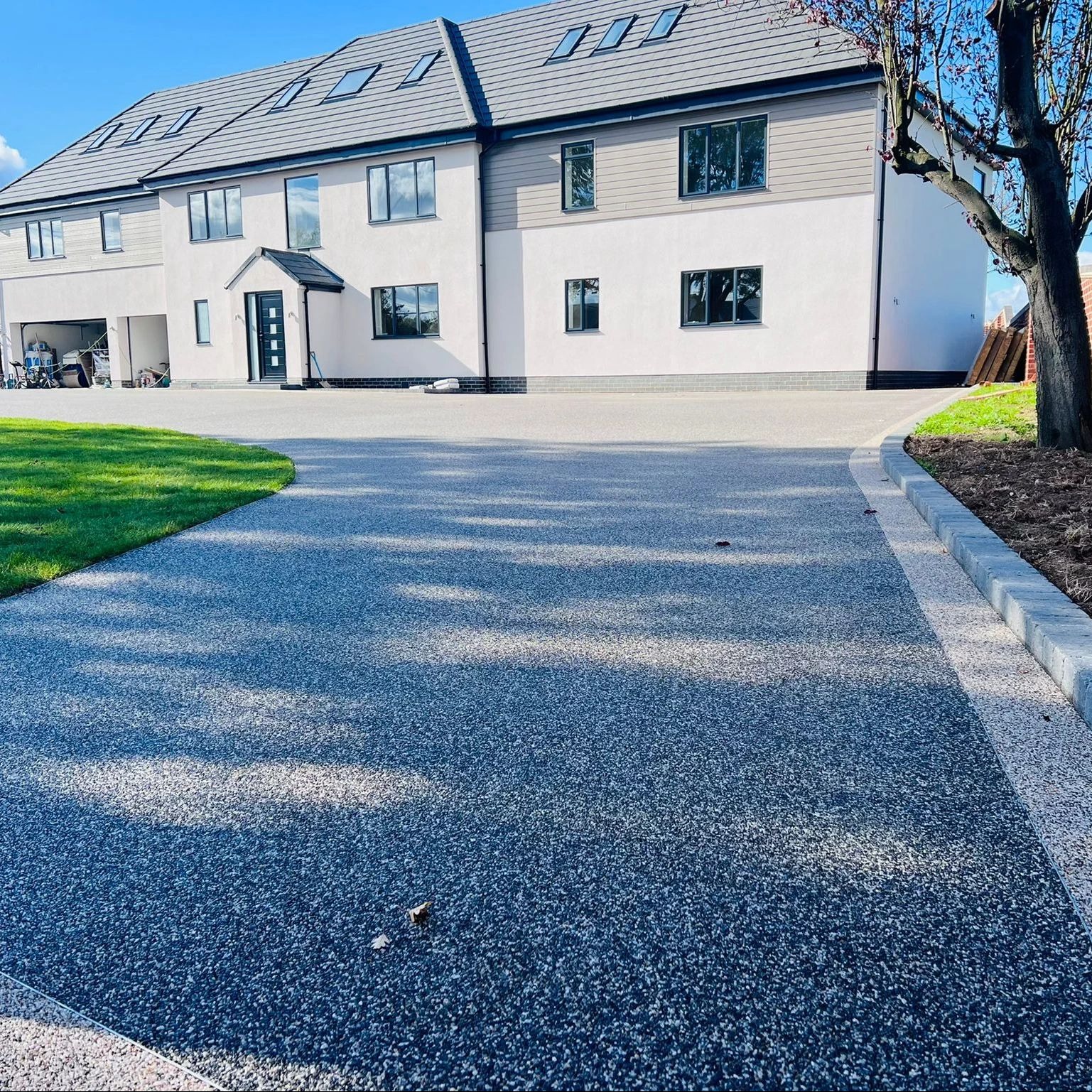 Nowoczesny podjazd z żywicy epoksydowej w odcieniach szarości, prowadzący do domu z garażem. Widoczne obrzeża i trawnik. Słoneczny dzień.
