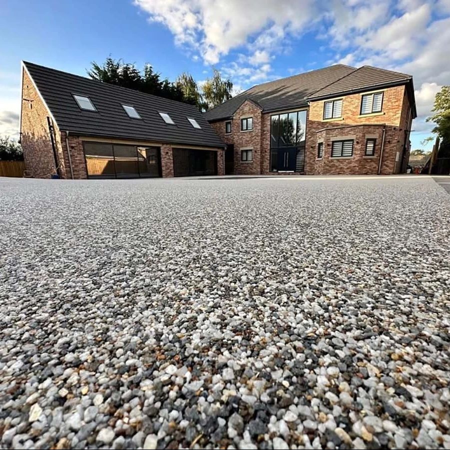 Nowoczesny dom z podjazdem wyłożonym drobnym, jasnym kamieniem. Ujęcie z niskiej perspektywy podkreśla fakturę kamienia i bryłę budynku.