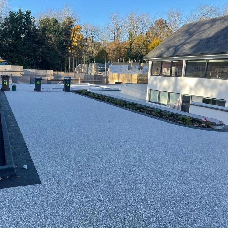 Jasnoszary taras żywiczny z niską roślinnością, widok na budynek z oknami i konstrukcję w tle, słoneczny dzień.