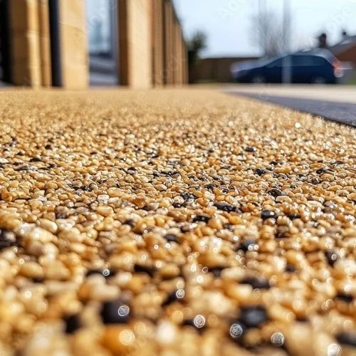 Żwirowa nawierzchnia tarasu w ciepłych odcieniach beżu i brązu, z widoczną fakturą drobnych kamyczków i subtelnym połyskiem, w tle budynek i samochód.