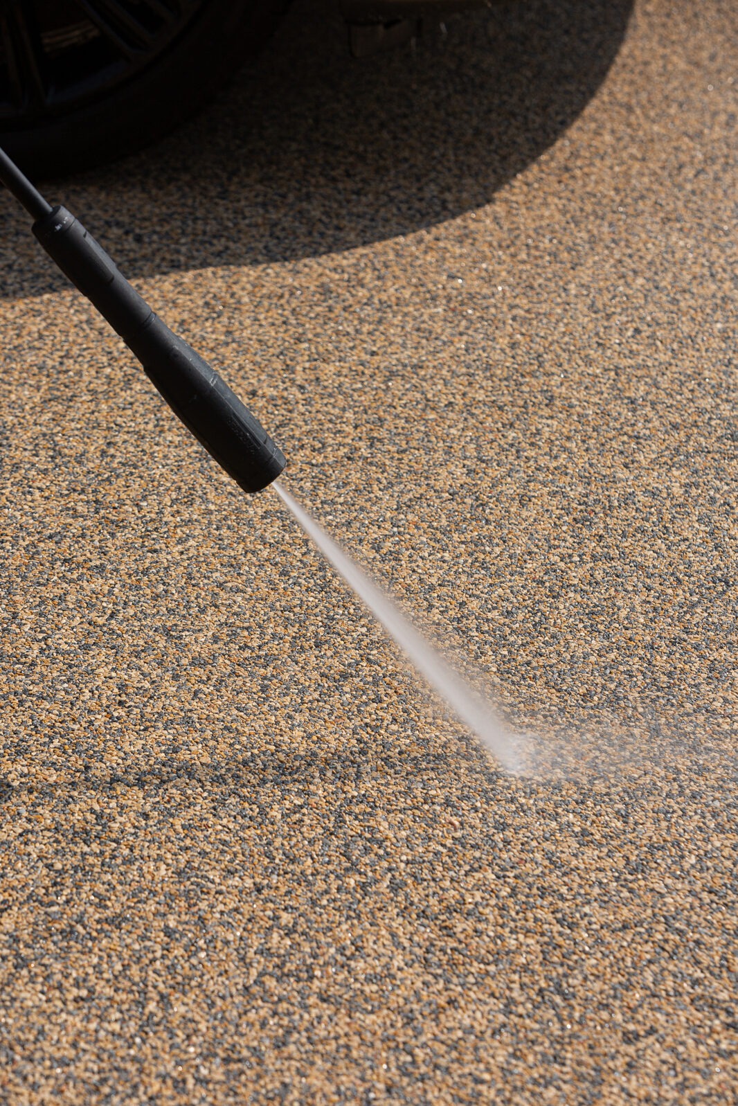 Czyszczenie powierzchni z kamyczków za pomocą myjki ciśnieniowej. Woda pod ciśnieniem usuwa zabrudzenia, ukazując naturalny kolor kamienia. Fragment opony w tle.