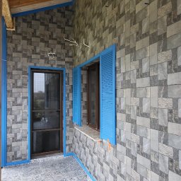 Tetproject - Zadaszony taras z mozaikową podłogą i ścianami wyłożonymi płytkami imitującymi kamień. Niebieskie ramy okienne i drzwiowe. Widoczne drewniane belki stropowe.