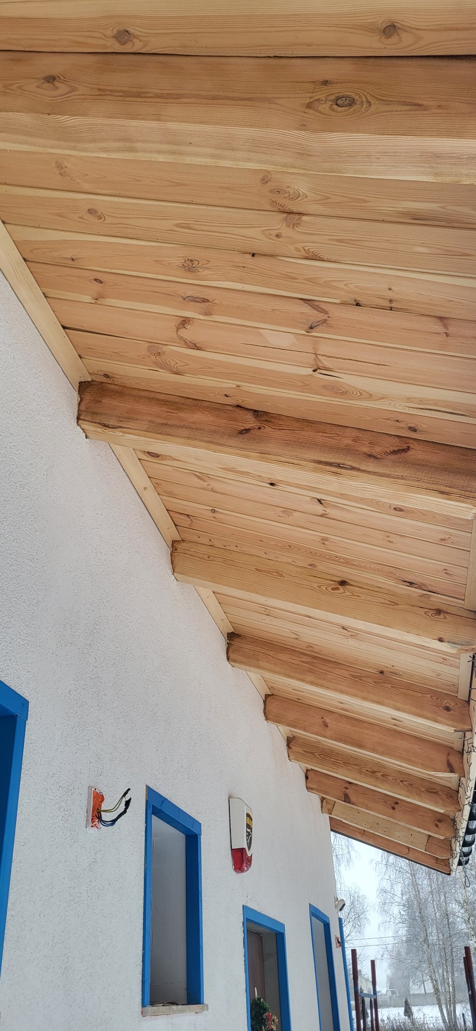 Drewniany podbitka dachowa z widocznymi belkami, biała elewacja z niebieskimi ramami okiennymi. Widok z dołu, zima w tle.