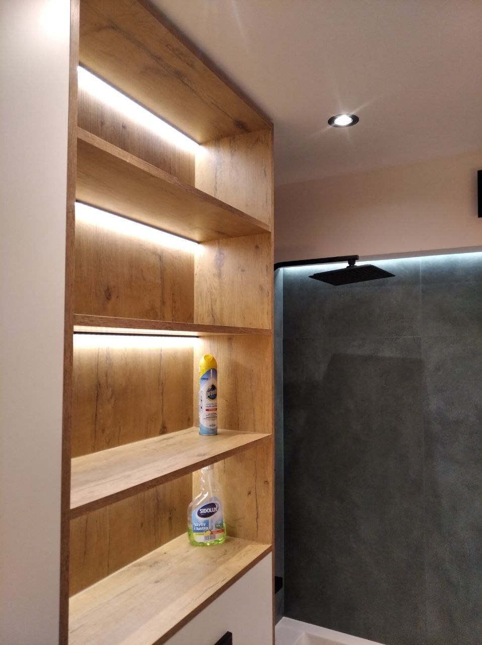 Nowoczesna łazienka z drewnianym regałem podświetlanym LED, szara ściana prysznicowa i czarna deszczownica. Minimalistyczny design.