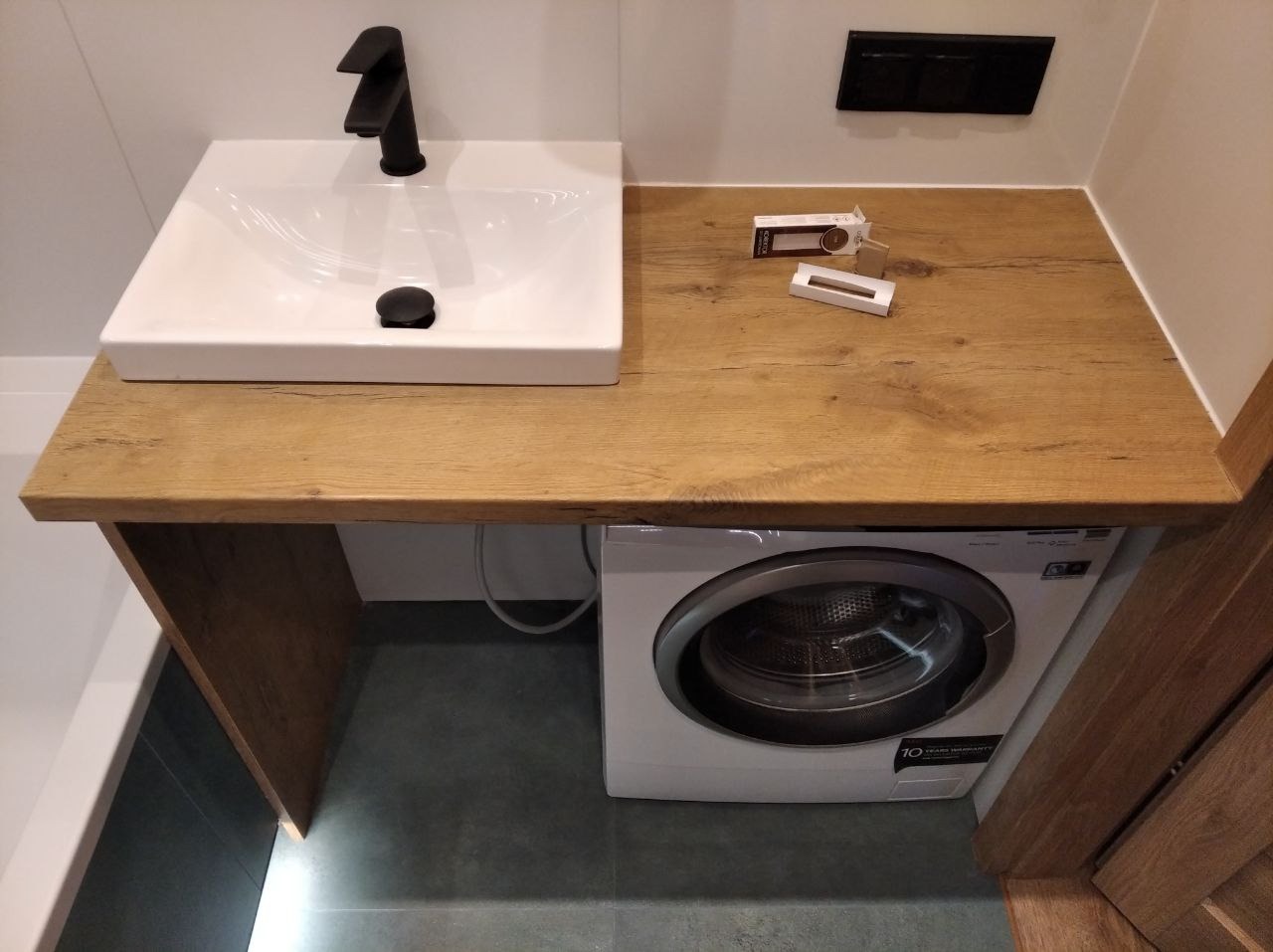 Nowoczesna łazienka z umywalką nablatową, czarną armaturą i blatem z efektem drewna, pod którym umieszczono pralkę. Minimalistyczny design.