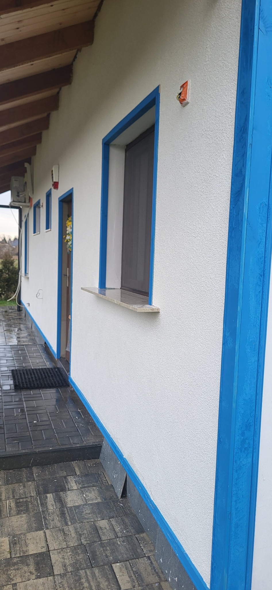 Biała elewacja domu z niebieskimi akcentami okien i narożników. Widoczny fragment chodnika z kostki brukowej i drewniane zadaszenie. Ujęcie z boku budynku.
