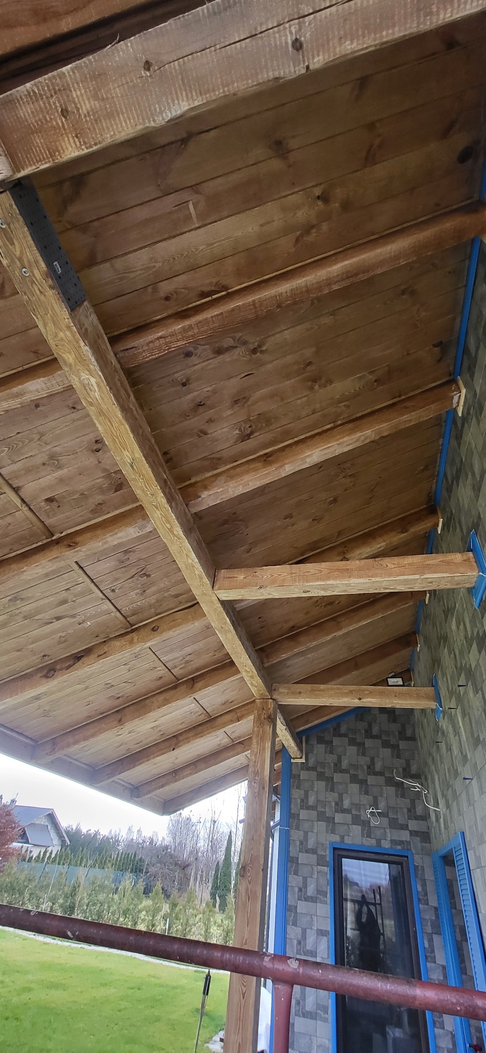 Drewniany strop z belek, widok z dołu, z fragmentem ściany wykończonej płytkami. Elementy konstrukcyjne połączone metalowymi wspornikami. Widoczne detale wykończenia.