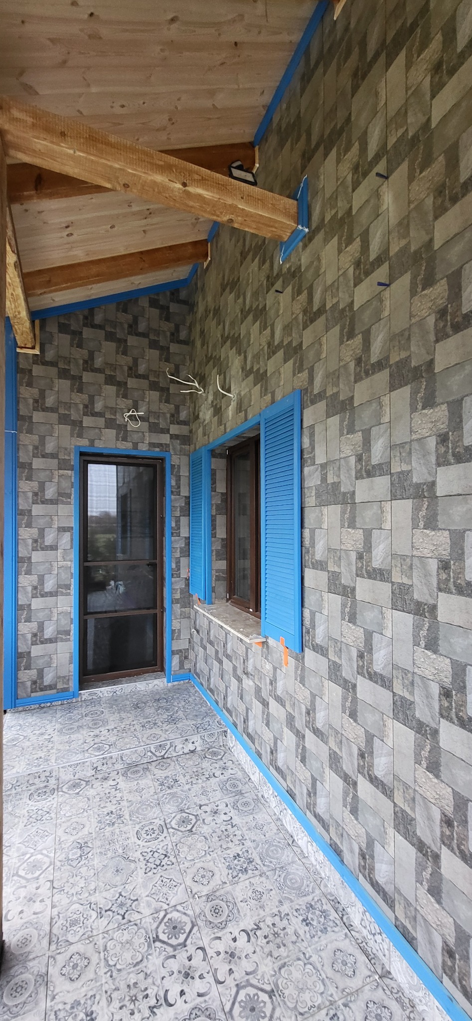 Zadaszony taras z mozaikową podłogą i ścianami wyłożonymi płytkami imitującymi kamień. Niebieskie ramy okienne i drzwiowe. Widoczne drewniane belki stropowe.