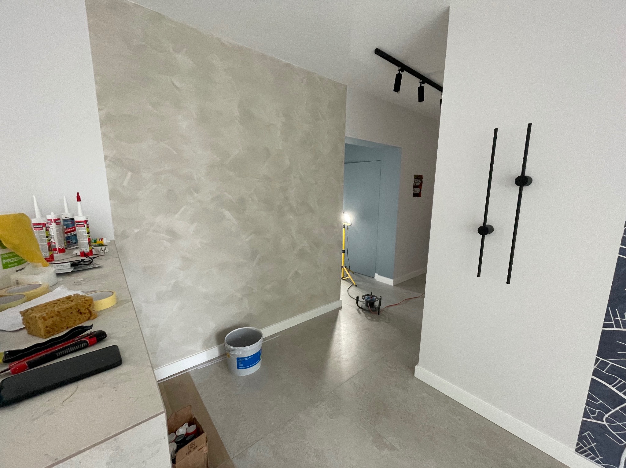 Ściana z efektem dekoracyjnym w odcieniach szarości, widok na korytarz z oświetleniem punktowym i narzędzia malarskie na blacie. Adaptacja biura w Gdyni.
