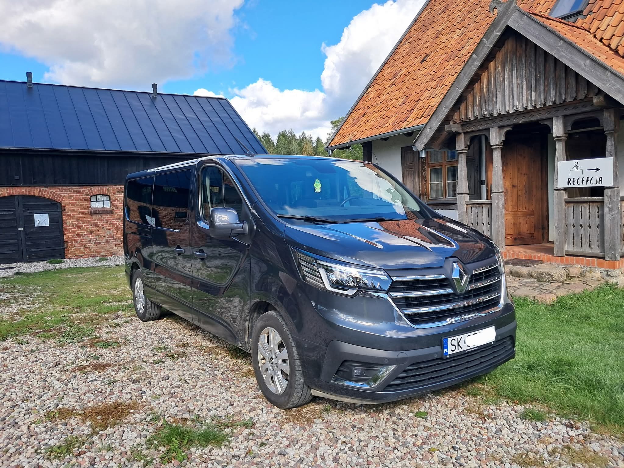 Ciemny bus Renault Trafic na żwirowym podjeździe przed budynkiem z drewnianą werandą i szyldem 'Recepcja'. Błękitne niebo z chmurami w tle.
