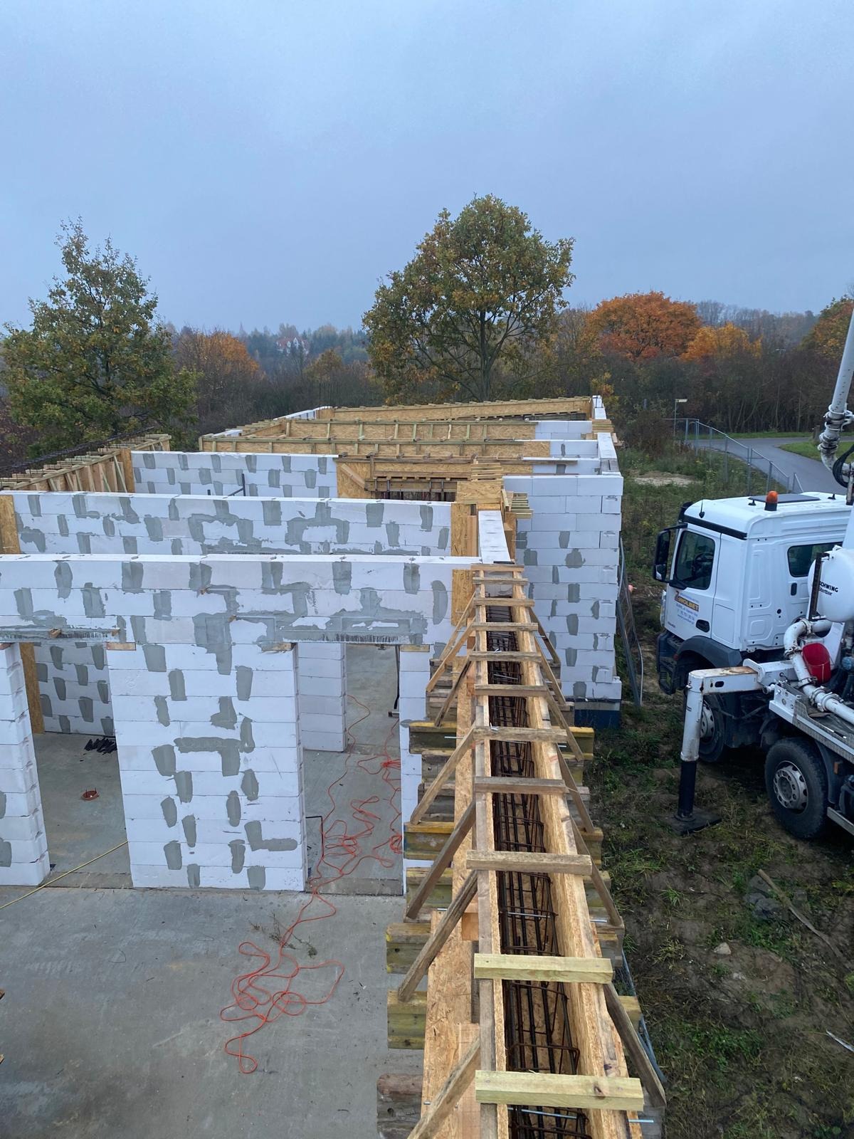 Wysoki kąt: Budowa domu z białych bloczków, widoczne drewniane szalunki zbrojenia, pompa do betonu i jesienne drzewa w tle. Etap wznoszenia ścian.