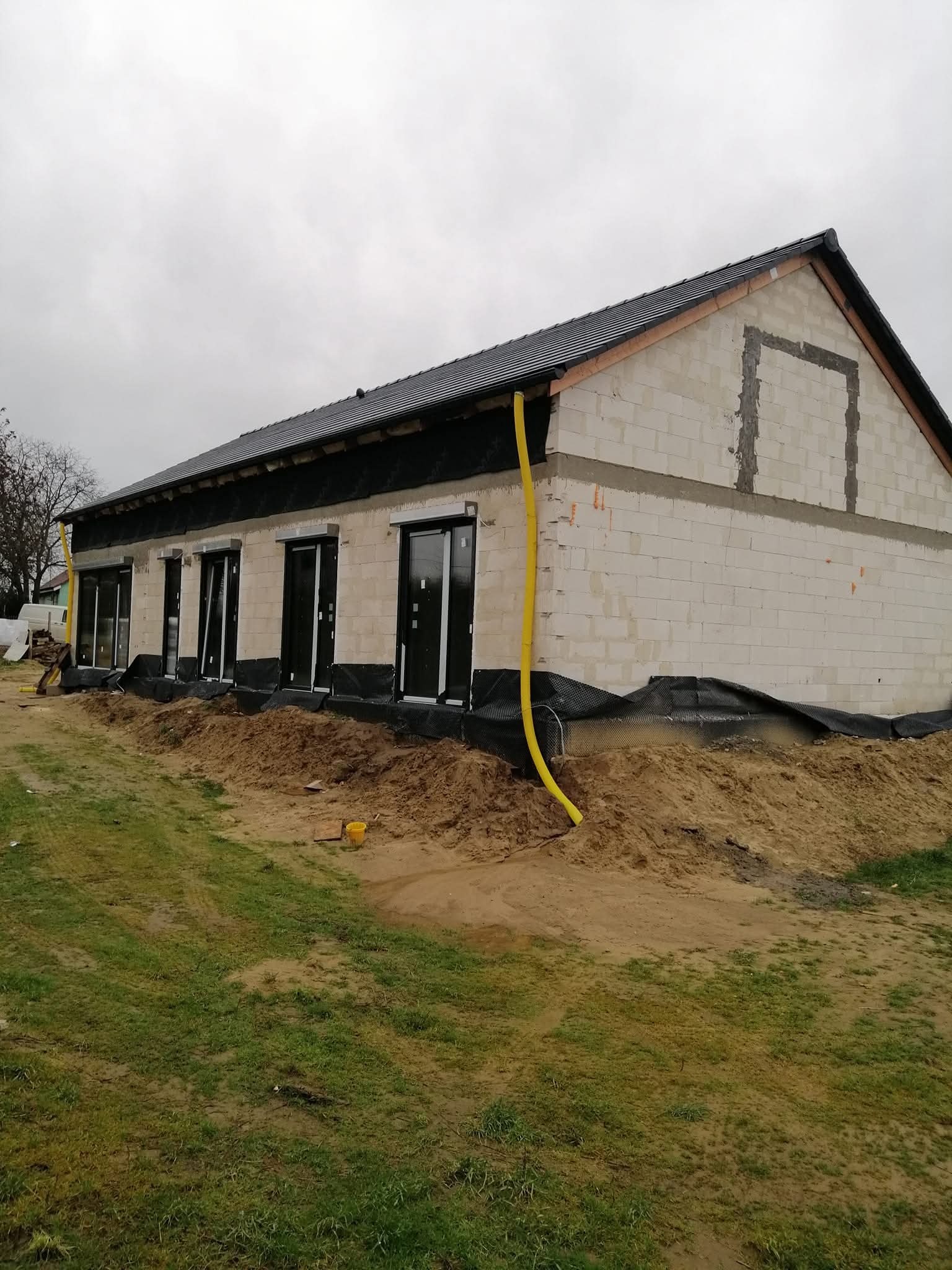 Niedokończony dom z białego pustaka z czarną membraną, widoczne okna i żółta rura gazowa. Etap budowy, szary dzień.