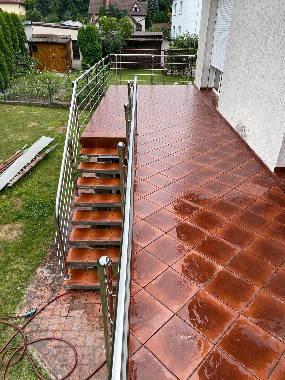 Błyszczący, brązowy taras z płytkami ułożonymi w karo, z metalową balustradą i schodami. Widok na ogród i budynki w tle. Świeżo po deszczu.