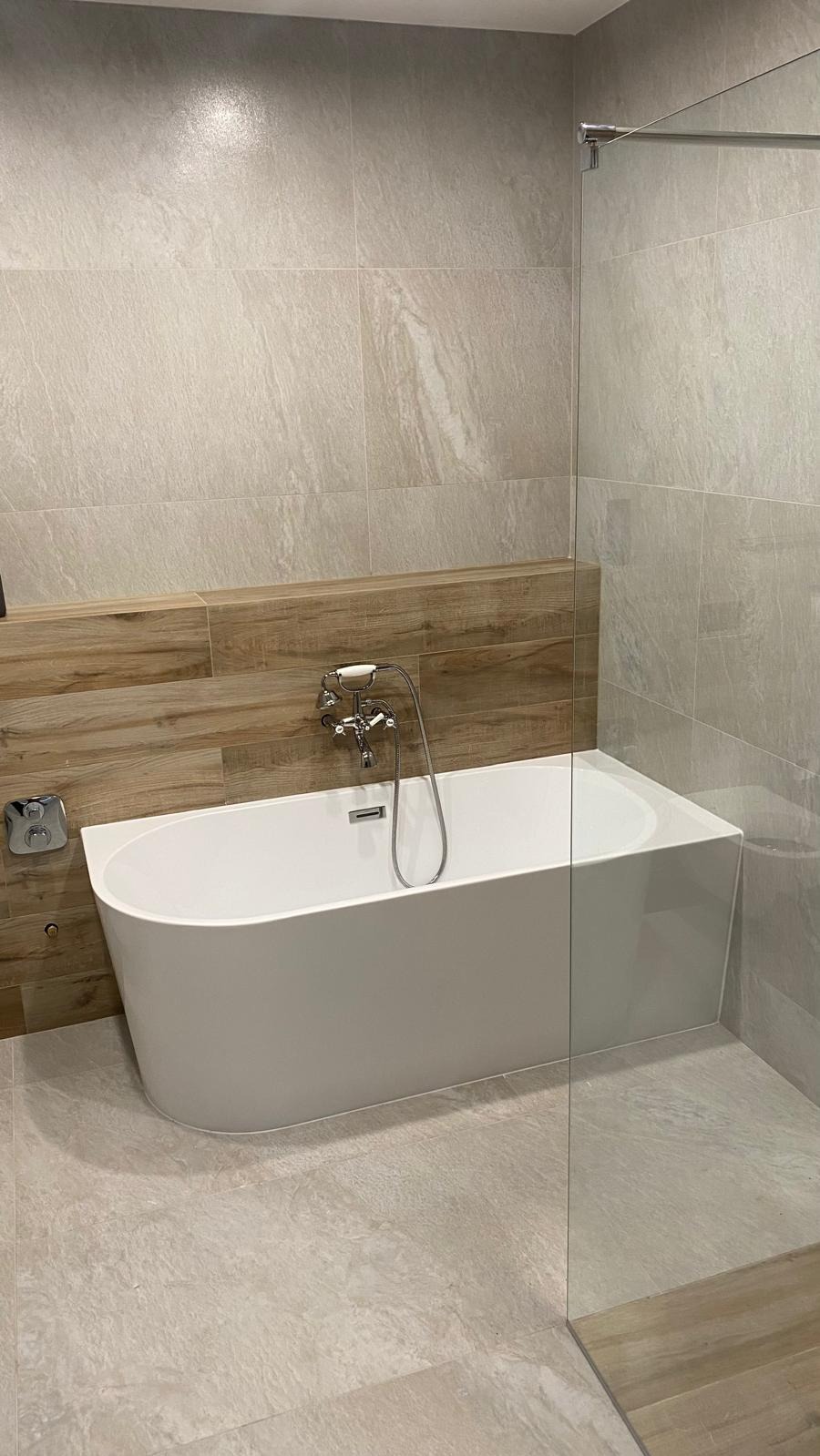 Nowoczesna łazienka z wolnostojącą wanną, szklaną kabiną i beżowymi płytkami imitującymi kamień. Drewniane akcenty nadają ciepła. Bateria wannowa retro.