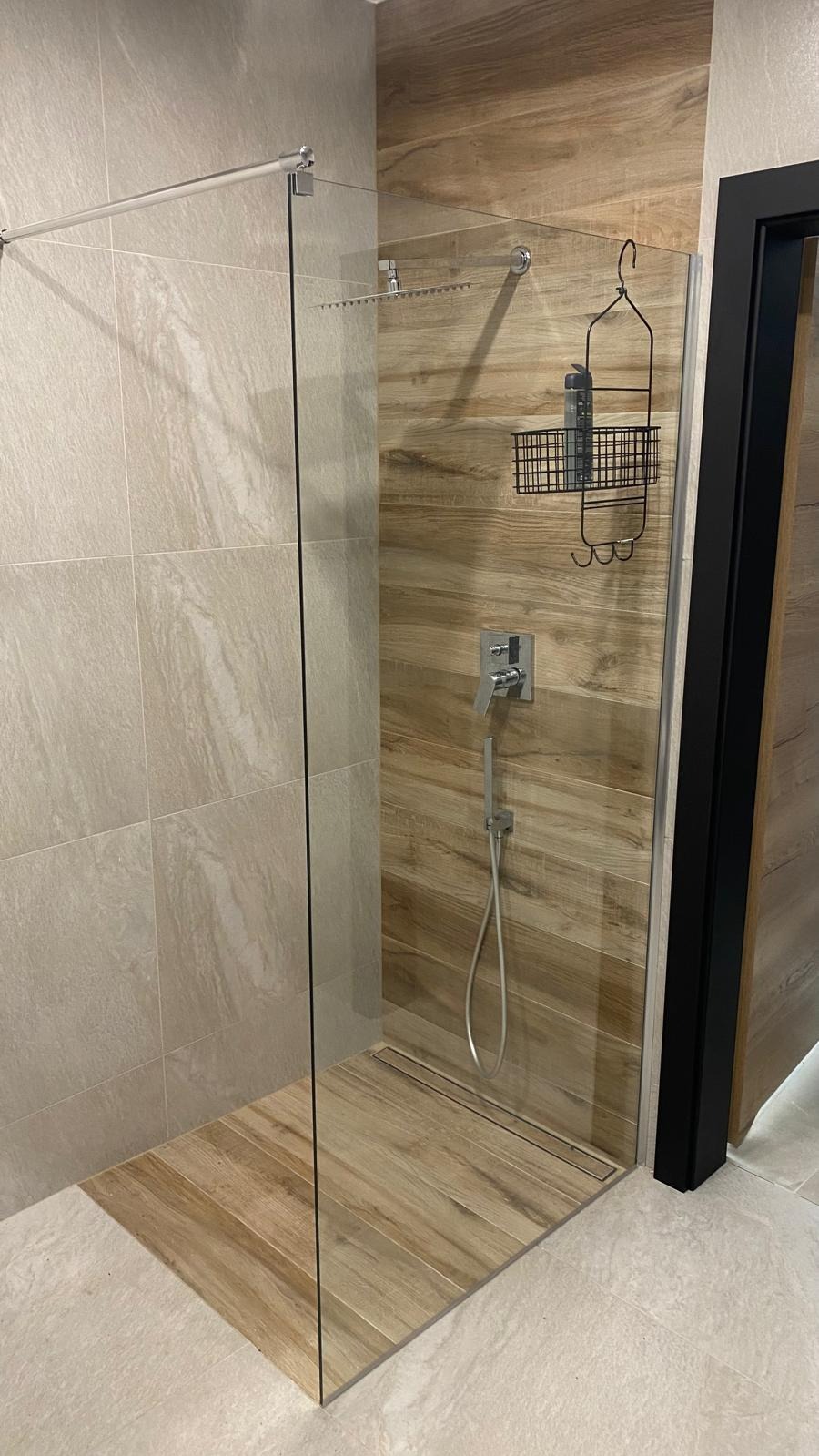 Nowoczesna kabina prysznicowa walk-in z drewnopodobnymi płytkami, chromowaną armaturą i czarnymi akcentami. Minimalistyczny design z odpływem liniowym.