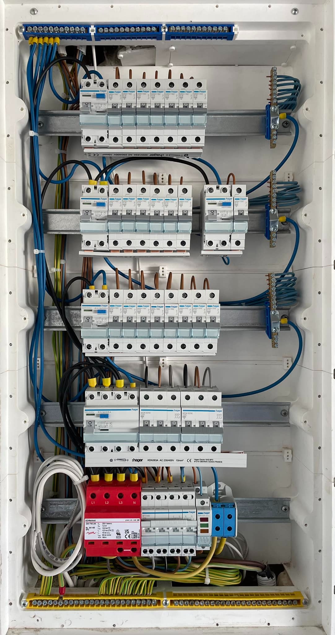 Rozdzielnica elektryczna Hager z zabezpieczeniami, przewodami i listwami zaciskowymi w białej obudowie. Widoczne bezpieczniki, ograniczniki przepięć i kable w różnych kolorach.