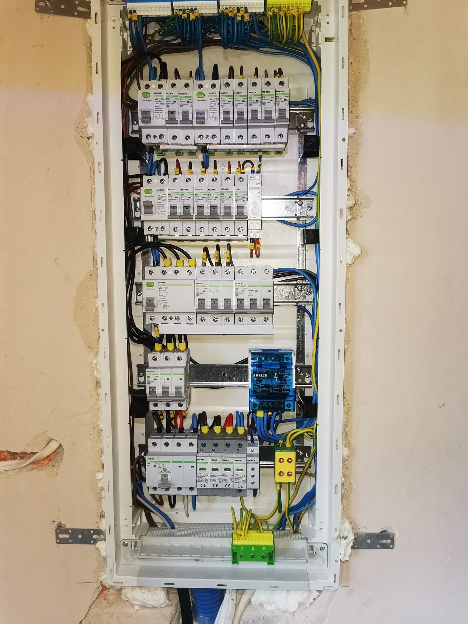Rozdzielnica elektryczna NOARK w trakcie montażu, widoczne zabezpieczenia, przewody w kolorach: niebieskim, żółto-zielonym, brązowym i czarnym. Ściana w tle z widocznymi śladami montażu.