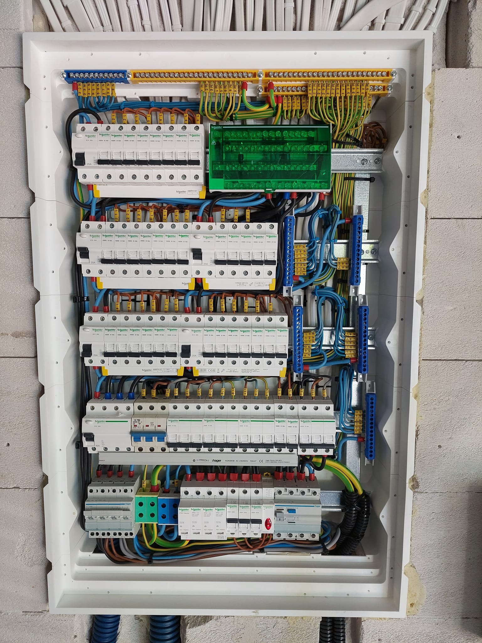 Rozdzielnica elektryczna w białej obudowie, z licznymi bezpiecznikami i przewodami oznaczonymi kolorami, zamontowana na ścianie z betonu komórkowego.