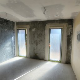 Fbu Srebniak - Pokój w trakcie prac wykończeniowych: ściany z nałożoną gładzią, zabezpieczone okna folią, surowy beton na podłodze, widoczne ślady szpachlowania i przygotowania do malowania.
