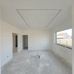 Fbu Srebniak - Wnętrze w trakcie prac wykończeniowych: białe ściany, sufit z ramą, surowa podłoga. Widoczne instalacje i okno z widokiem na budynek. Jasne, naturalne światło.