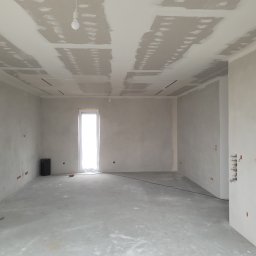 Fbu Srebniak - Pokój w trakcie prac wykończeniowych: ściany i sufit pokryte gładzią, widoczne łączenia płyt karton-gips, surowe instalacje elektryczne. Okno z białymi ramami.