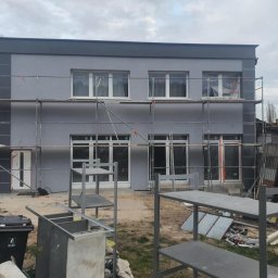 Sebastian Jan - Szara elewacja domu jednorodzinnego w trakcie prac wykończeniowych. Widoczne rusztowania, okna zabezpieczone folią, metalowe regały i śmietnik na pierwszym planie.
