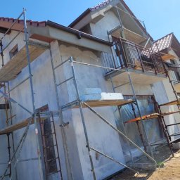 Sebastian Jan - Dom w trakcie tynkowania elewacji, widoczne rusztowania, zabezpieczone okna folią. Świeży tynk w jasnym odcieniu. Częściowo wykończony balkon z metalową balustradą.